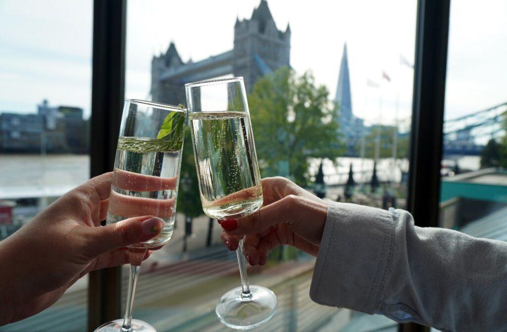 Two people clinking glasses of sparkling wine beside a window, with Tower Bridge and the London skyline visible in the background.