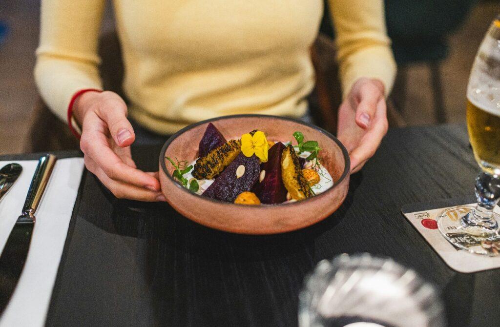 Diner holding a colourful bowl of roasted vegetables garnished with greens and an edible flower at a restaurant table, with a glass of beer alongside.