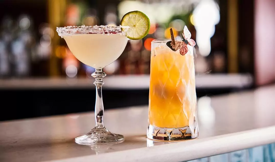 A margarita with a lime slice and salted rim next to a tall iced cocktail on a bar counter.
