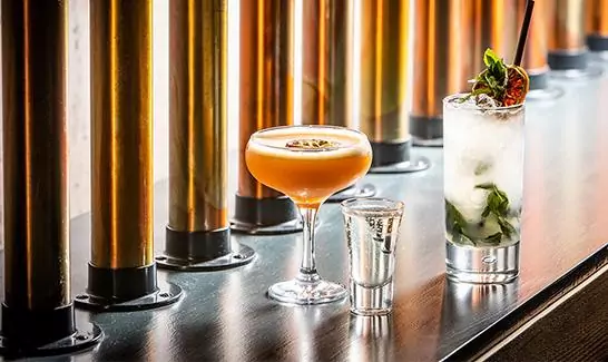 Three drinks on a bar counter: a cocktail in a coupe glass, a shot in a small glass, and a mojito garnished with mint and a lime slice.