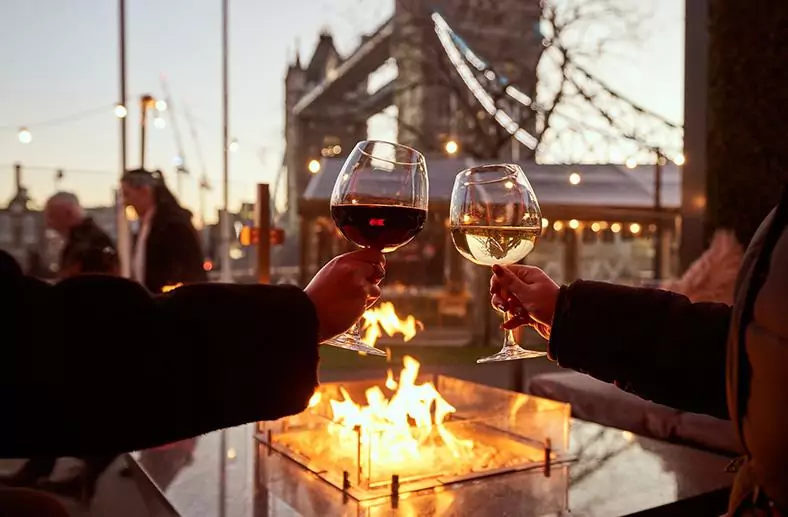 Two individuals clink wine glasses together, celebrating over a warm fire pit in a cozy outdoor setting with tower bridge in the background.