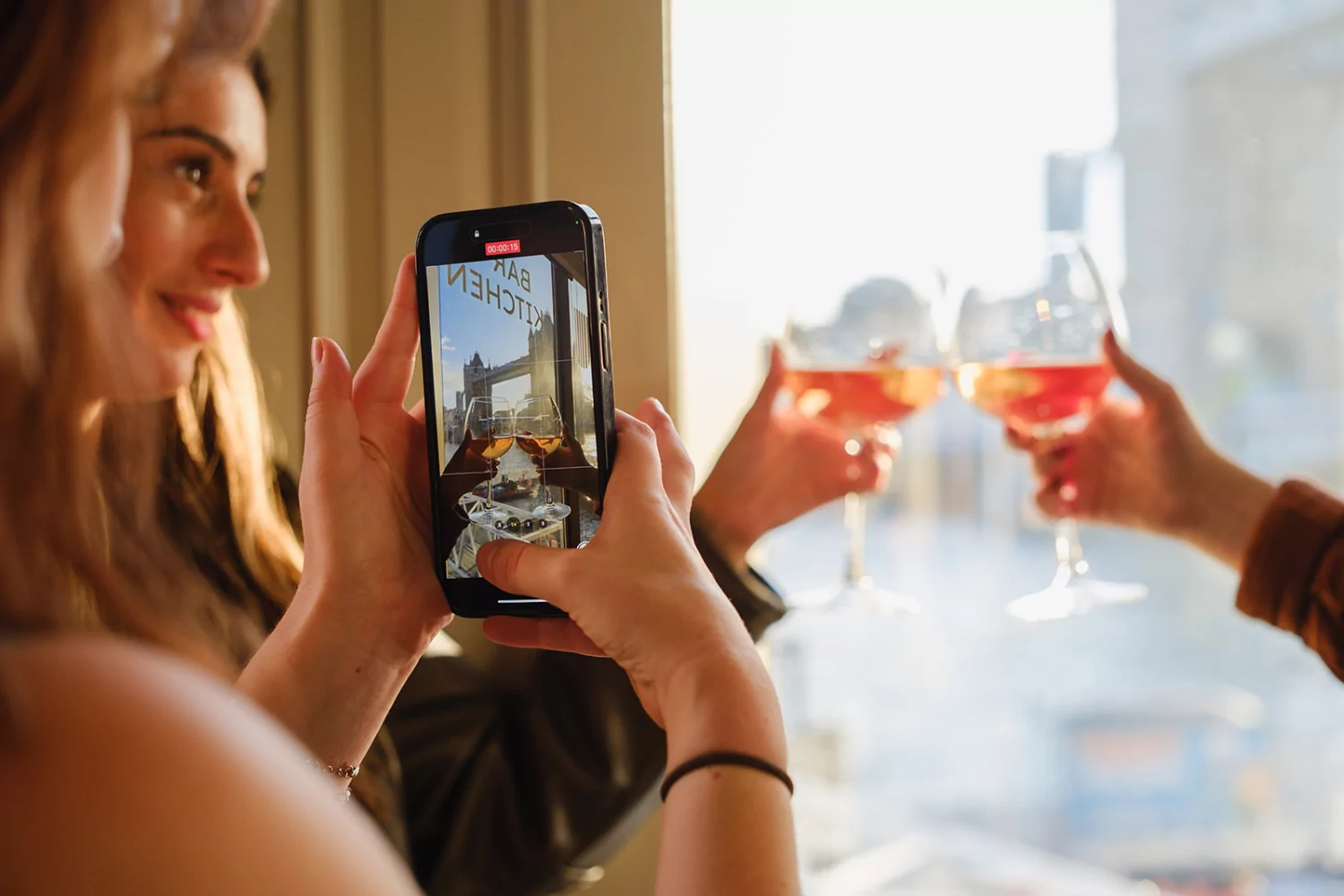 Person holding a smartphone to photograph two glasses of rosé being clinked together by a window, with Tower Bridge visible in the background.